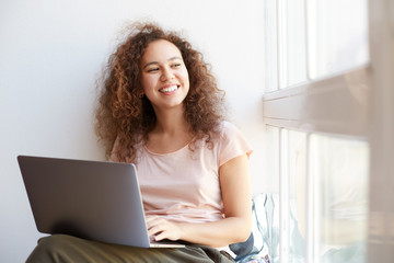 Portrait of cheerful young curly african american girl sitting by the window, working at laptop and enjoying freelance work and sunny day at home.