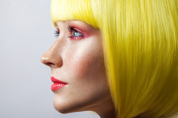 Beauty side view profile portrait of cute young calm female model with freckles, red makeup and yellow wig, looking forward with serious face. indoor studio shot, isolated on gray background.