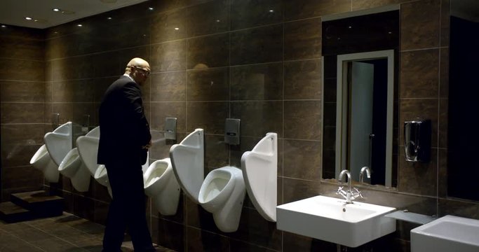 A bald man in glasses in a business suit in the bathroom near the urinal, zips his pants and jacket and goes to wash his hands in the sink at the mirror, stroking his bald head.