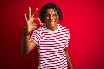 Young afro man with dreadlocks wearing striped t-shirt standing over isolated red background smiling positive doing ok sign with hand and fingers. Successful expression.