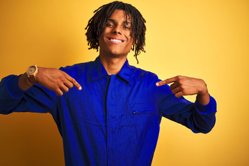 Afro worker man with dreadlocks wearing mechanic uniform over isolated yellow background looking confident with smile on face, pointing oneself with fingers proud and happy.