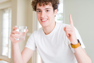 Young man drinking a glass of water at home surprised with an idea or question pointing finger with happy face, number one