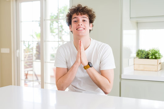 Young handsome man wearing white t-shirt at home praying with hands together asking for forgiveness smiling confident.