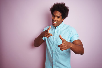 Young american man with afro hair wearing blue shirt standing over isolated pink background pointing fingers to camera with happy and funny face. Good energy and vibes.