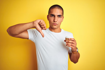 Young handsome man drinking a take away glass of coffee over yellow isolated background with angry face, negative sign showing dislike with thumbs down, rejection concept