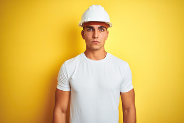 Young handsome man wearing construction helmet over yellow isolated background with serious expression on face. Simple and natural looking at the camera.