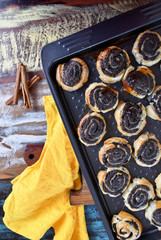 Top view: an image of a baking tray with baked rolls with poppy seeds on a cutting board, three sticks of cinnamon and a yellow kitchen napkin on a multi-colored table