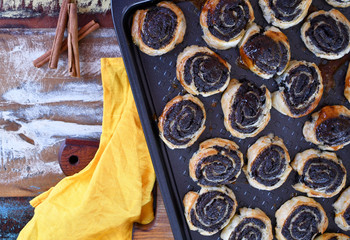 Top view: an image of a baking tray with baked rolls with poppy seeds on a cutting board, three sticks of cinnamon and a yellow kitchen napkin on a multi-colored table