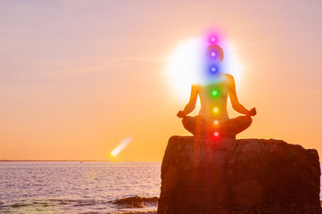 Woman is meditating with glowing seven chakras on stone at sunset. Silhouette of woman is...