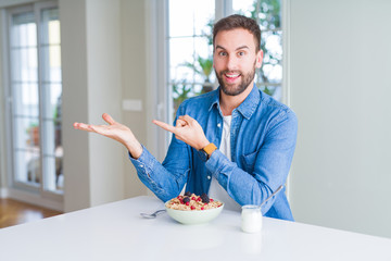 Handsome man eating cereals for breakfast at home amazed and smiling to the camera while presenting with hand and pointing with finger.