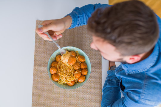 Handsome man eating pasta with meatballs and tomato sauce at home while smiling at the camera
