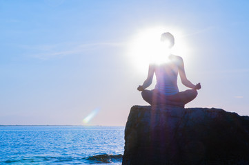 Woman is practicing yoga sitting on stone in Lotus pose at sunset. Silhouette of woman meditating on the beach