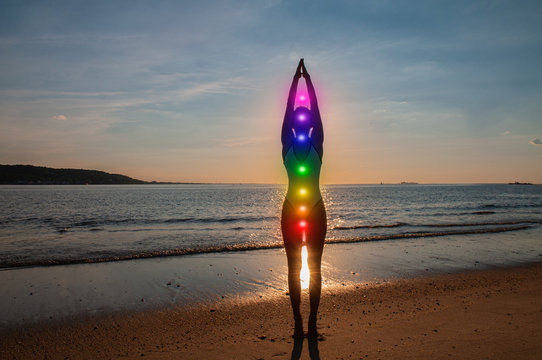 Woman is meditating with glowing seven chakras on the beach. Silhouette of woman is practicing yoga at sunset.