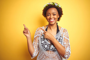 Young african american woman with afro hair wearing flowers crown over yellow isolated background smiling and looking at the camera pointing with two hands and fingers to the side.