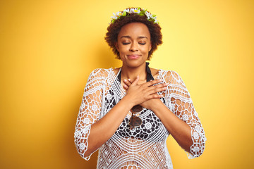 Young african american woman with afro hair wearing flowers crown over yellow isolated background smiling with hands on chest with closed eyes and grateful gesture on face. Health concept.