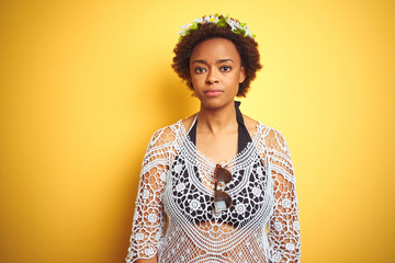 Young african american woman with afro hair wearing flowers crown over yellow isolated background with serious expression on face. Simple and natural looking at the camera.
