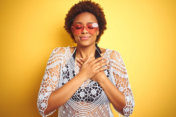Young african american woman with afro hair wearing bikini and heart shaped sunglasses smiling with hands on chest with closed eyes and grateful gesture on face. Health concept.