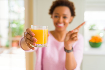 Young african american woman driking orange juice at home very happy pointing with hand and finger to the side