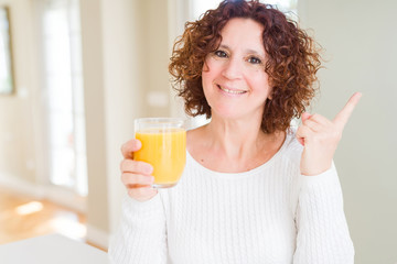 Senior woman driking a glass of fresh orange juice very happy pointing with hand and finger to the side