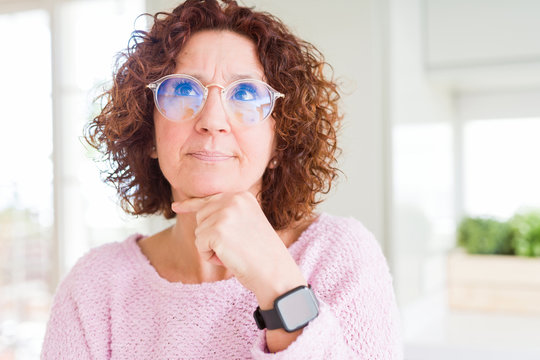 Beautiful senior woman wearing pink sweater and glasses with hand on chin thinking about question, pensive expression. Smiling with thoughtful face. Doubt concept.