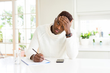 African american student man writing on a paper using a pencil stressed with hand on head, shocked with shame and surprise face, angry and frustrated. Fear and upset for mistake.