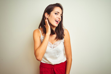 Young beautiful woman wearing t-shirt standing over white isolated background smiling with hand over ear listening an hearing to rumor or gossip. Deafness concept.