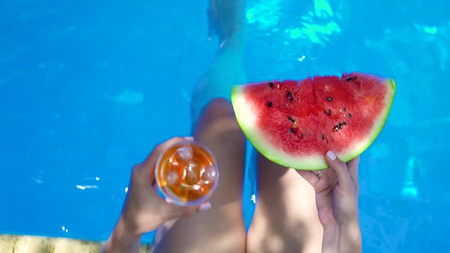 Woman moves her legs in the pool with a slice of watermelon and a glass of aperol
