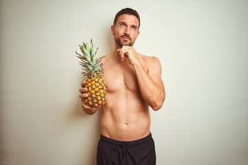 Handsome shirtless man wearing swimwear and holding pineapple isolated background serious face...