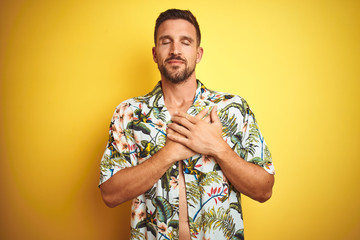 Handsome man wearing summer hawaiian flowers shirt over yellow isolated background smiling with hands on chest with closed eyes and grateful gesture on face. Health concept.