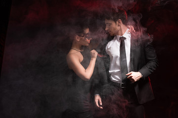 selective focus of sexy woman in mask undressing man in smoke on black