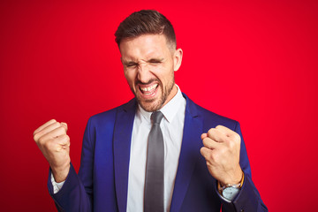 Young handsome business man over red isolated background very happy and excited doing winner gesture with arms raised, smiling and screaming for success. Celebration concept.