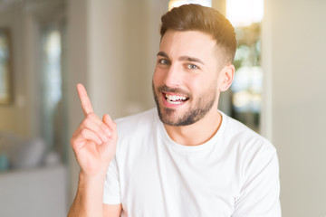 Young handsome man wearing casual white t-shirt at home with a big smile on face, pointing with hand and finger to the side looking at the camera.