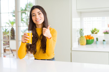 Young woman drinking a glass of fresh orange juice happy with big smile doing ok sign, thumb up with fingers, excellent sign