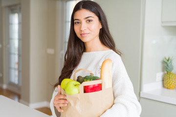 Beautiful young woman smiling holding a paper bag full of groceries at the kitchen