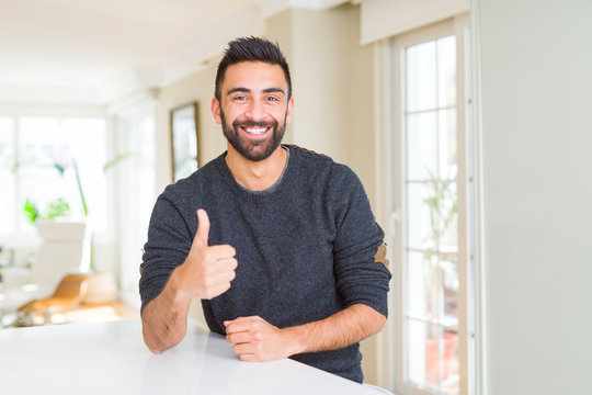 Handsome hispanic man wearing casual sweater at home doing happy thumbs up gesture with hand. Approving expression looking at the camera with showing success.