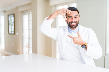 Handsome hispanic doctor or therapist man wearing medical coat at the clinic smiling making frame with hands and fingers with happy face. Creativity and photography concept.