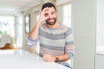 Handsome hispanic man wearing casual sweater at home doing ok gesture with hand smiling, eye looking through fingers with happy face.