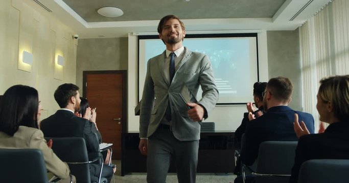 Cheerful and successful businessman speaker finishing his coaching speech at the conference and audience applauding him while he going away through people sitting in the rows.