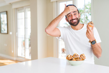 Handsome hispanic man eating chocolate chips muffin stressed with hand on head, shocked with shame and surprise face, angry and frustrated. Fear and upset for mistake.