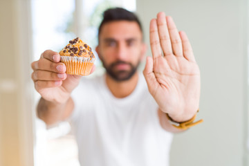 Handsome hispanic man eating chocolate chips muffin with open hand doing stop sign with serious and...