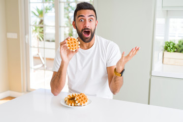 Handsome hispanic man eating sweet belgian waffle pastry very happy and excited, winner expression celebrating victory screaming with big smile and raised hands