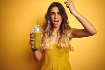 Young beautiful woman drinking detox green smoothy over yellow isolated background annoyed and frustrated shouting with anger, crazy and yelling with raised hand, anger concept