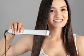 Young woman using hair iron on grey background