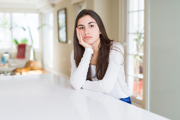 Beautiful young woman sitting on white table at home thinking looking tired and bored with...