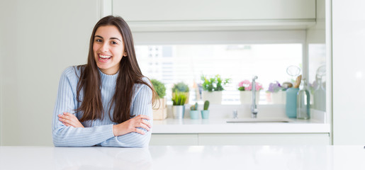 Wide angle picture of beautiful young woman sitting on white table at home happy face smiling with crossed arms looking at the camera. Positive person.