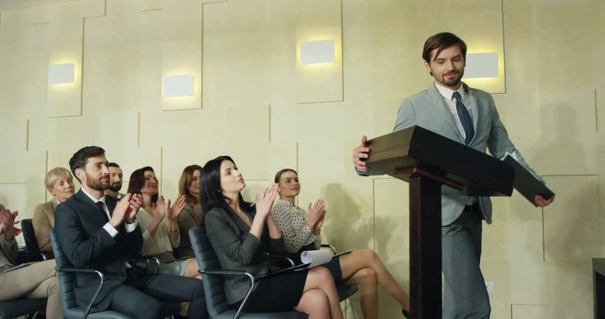 Caucasian young successful man business coach enterig the hall and going through the audience while people applauding, then coming to the lecturn and starting his educational speech.