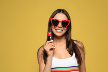 Fototapeta premium Portrait of beautiful young woman with heart shaped sunglasses and lollipop on color background