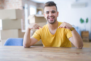 Young man sitting on the table with cardboard boxes behind him moving to new home looking confident with smile on face, pointing oneself with fingers proud and happy.