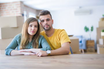 Obraz premium Young couple sitting on the table movinto to new home with carboard boxes behind them with serious expression on face. Simple and natural looking at the camera.