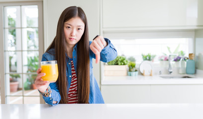 Beautiful Asian woman drinking a glass of fresh orange juice with angry face, negative sign showing dislike with thumbs down, rejection concept